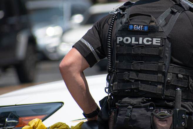 The image shows a police officer from the back, wearing a vest labeled "POLICE" and "EMT." This accompanies an article about excessive force in New York.